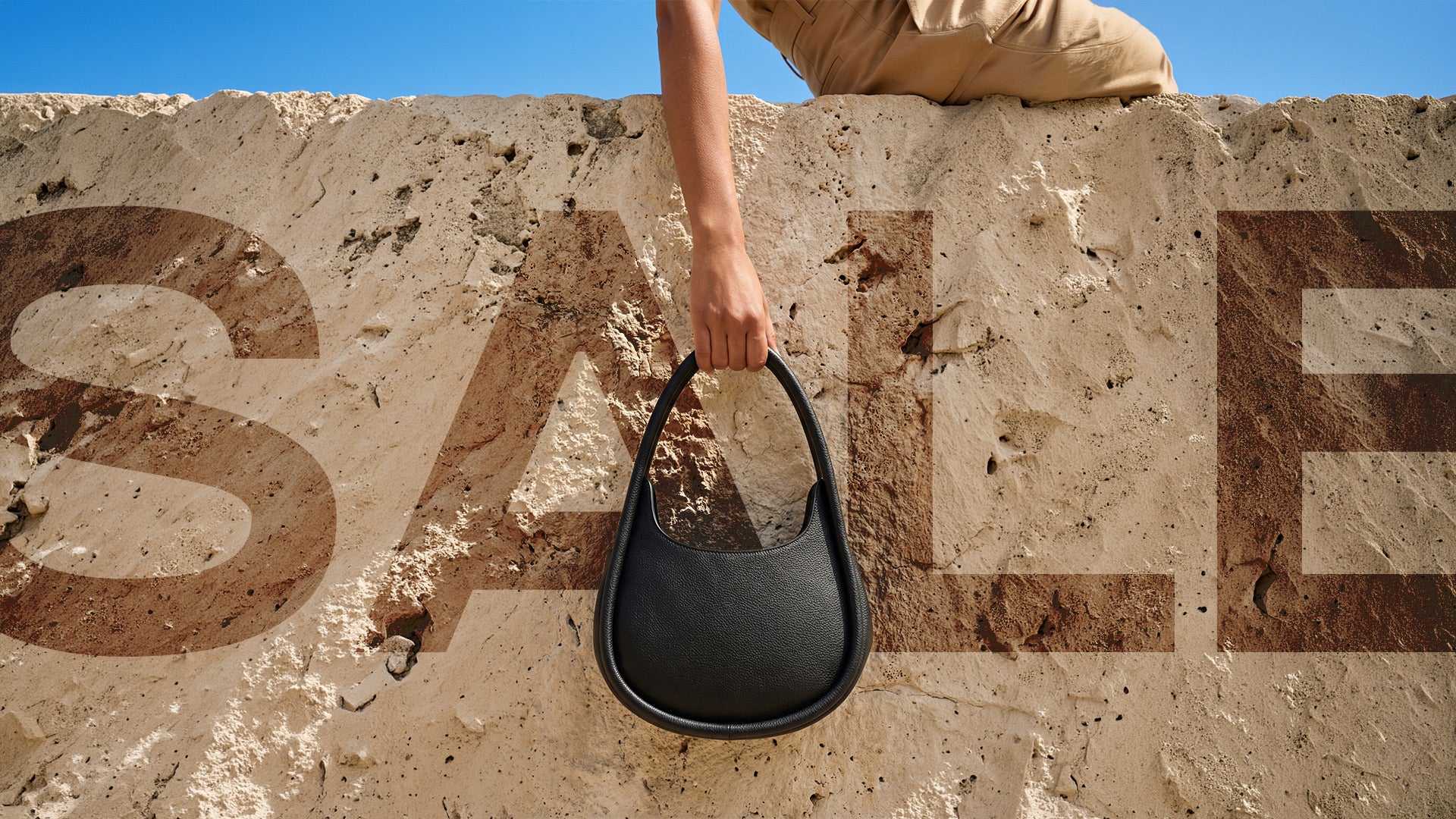 Person holding a black handbag against a stone wall with 'SALE' written on it.